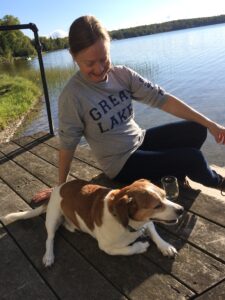 Jennifer Brunk (a white woman) and a beagle mix sit together on a lakeside dock. Jennifer is looking down at the dog and smiling 
