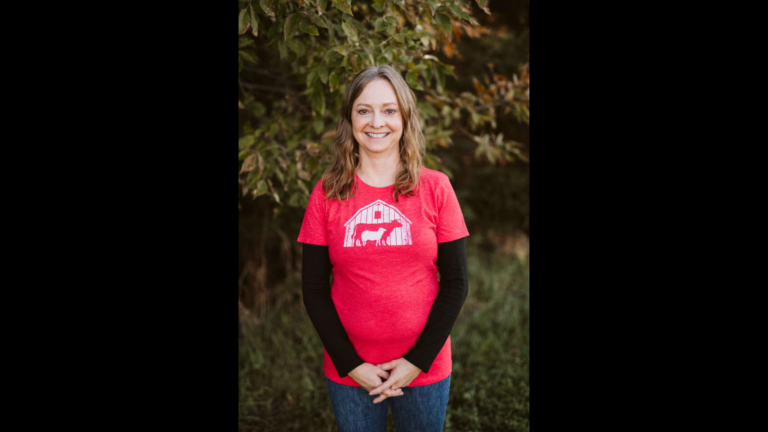 A photo of a white woman smiling in front of leafy foliage. She is wearing a red t-shirt with a design of a mother and baby cow.